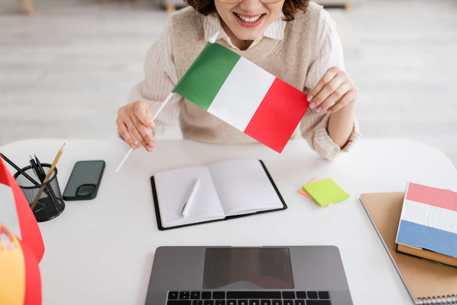 Cropped view of smiling language teacher holding Italian flag near devices and notebooks on table.
