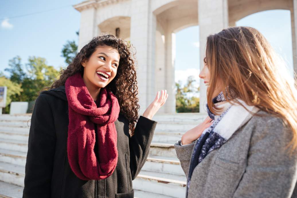 Two young women wearing scarves looking at each other and talking on a city street