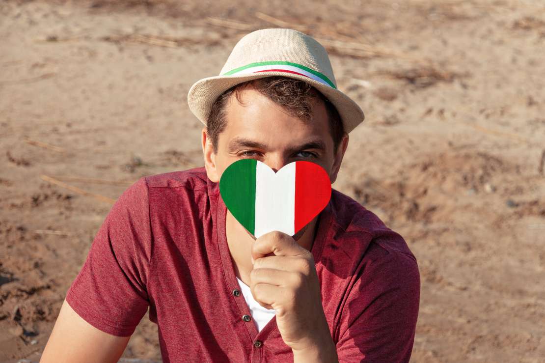 tourist smiling man in red t-shirt and hat covers face of wooden heart with italy flag